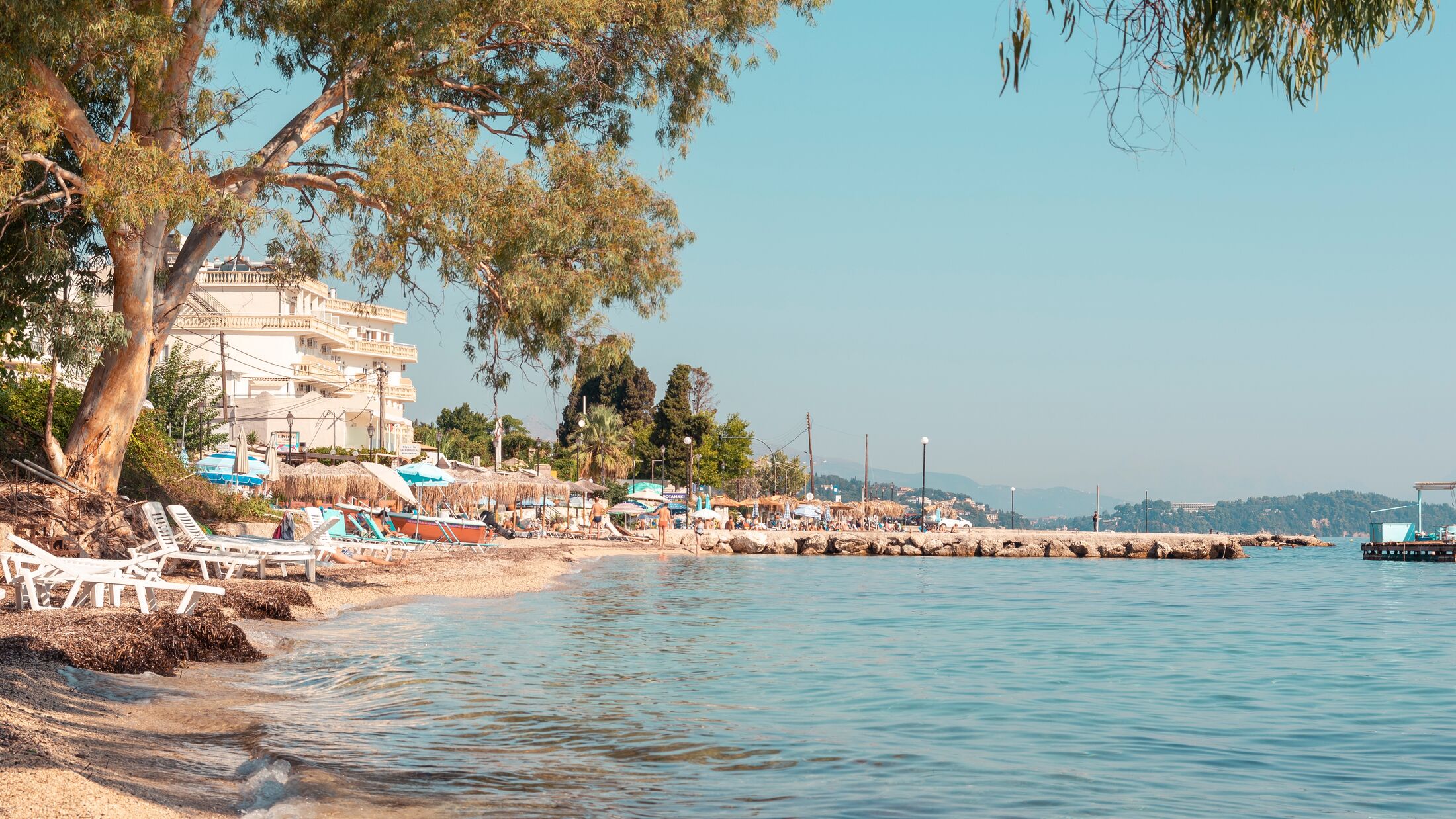Mediterranean beach view from a greek island, Corfu bay coastline in Greece on a sunny day, people having fun at seaside, colorful beach parasols and sun beds in front of the hotel